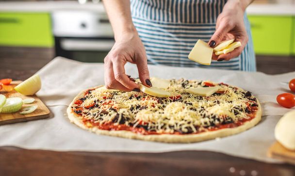 Una cocinera en delantal está poniendo queso mozzarella en una pizza cruda. Cocinar deliciosa pizza en casa en la cocina. - Foto, imagen
