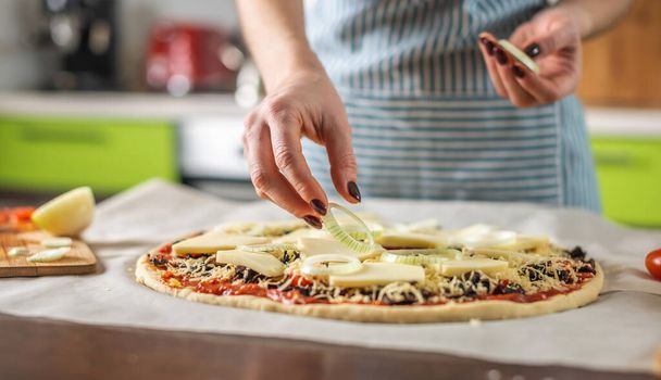 Een vrouwelijke chef in een schort doet uienringen om een rauwe pizza. Heerlijke pizza thuis koken in de keuken. - Foto, afbeelding