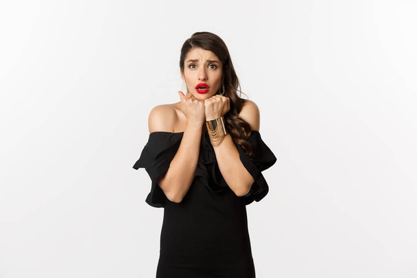 Fashion and beauty concept. Scared timid woman grimacing, looking concerned and worried at camera, standing over white background in black dress - Photo, Image