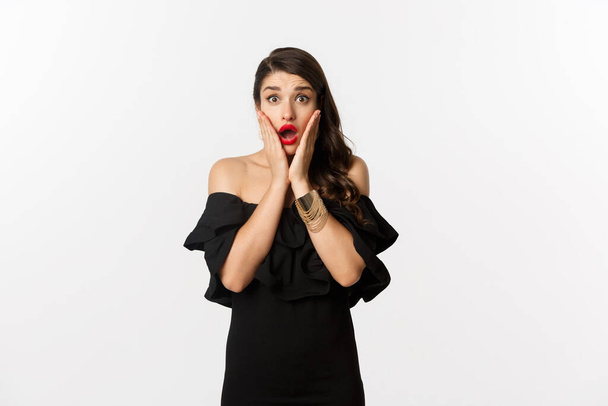 Beauty and fashion concept. Waist-up shot of surprised woman in black cocktail dress, looking amazed at camera, open mouth fascinated, standing over white background - Photo, Image