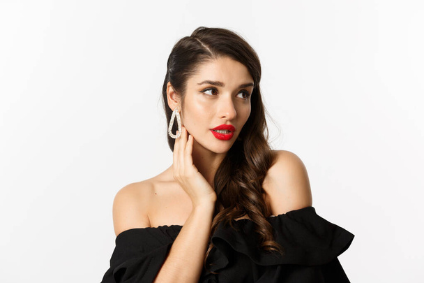 Mode en schoonheid concept. Close-up van elegante vrouw in zwarte jurk, met oorbellen en sensuele, rode lippenstift en make-up op, witte achtergrond - Foto, afbeelding