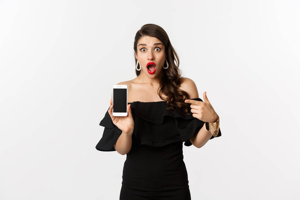 Online shopping concept. Fashionable woman in black dress pointing finger at smartphone screen, showing application, standing over white background - Foto, imagen