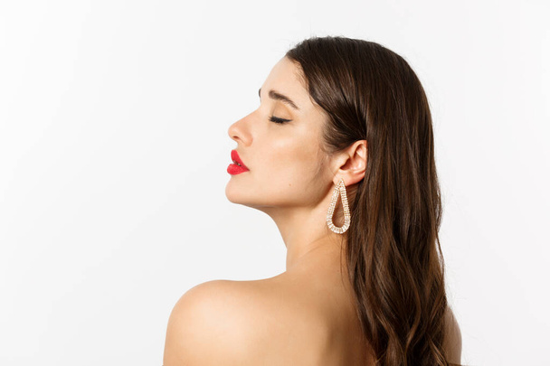 Fashion and beauty concept. Profile of beautiful woman standing naked with makeup and earrings, looking tender and sensual, standing over white background - Foto, Bild