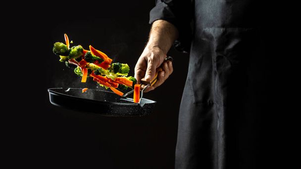 chef preparing food in the kitchen of a restaurant. flying vegetables scattering in a freeze motion of a cloud midair on black. Cook hands with vegetables on a pan. - Photo, Image