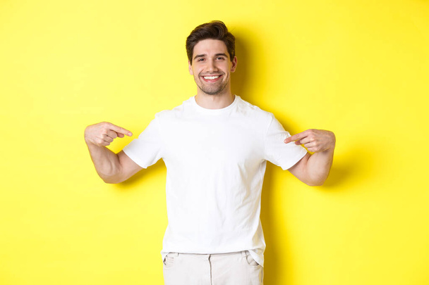 Parmakla logoyu gösteren, tişörtünde reklamını gösteren, sarı arka planda duran mutlu çekici adam. - Fotoğraf, Görsel