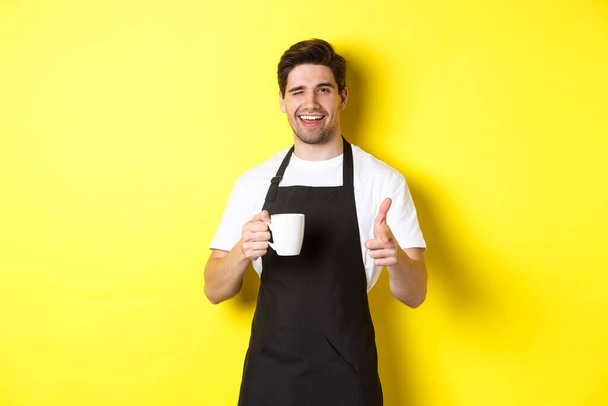 Barista guapo en delantal negro sosteniendo la taza de café, señalándote con el dedo, invitando a visitar la cafetería, de pie sobre el fondo amarillo - Foto, Imagen