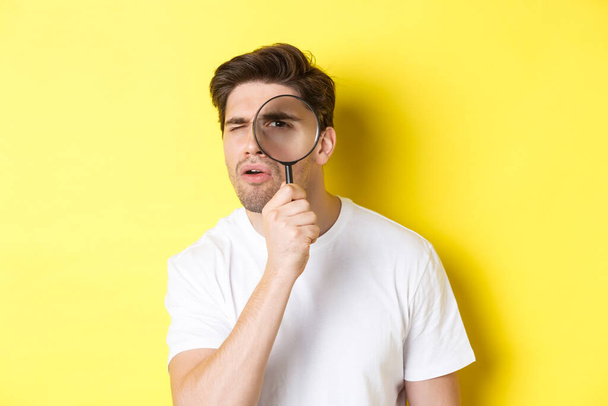 Man looking through magnifying glass with serious and thoughtful look, searching or investigating, standing over yellow background - Photo, Image