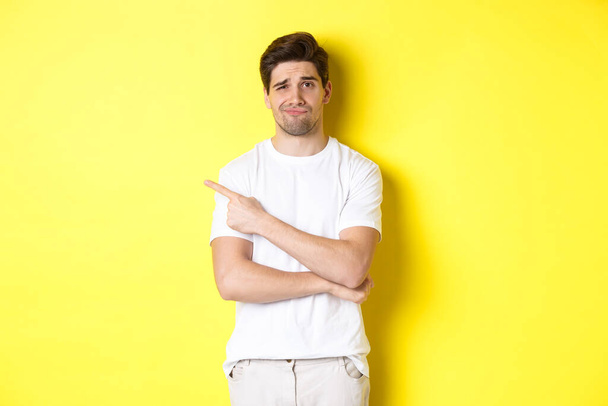 Скептически настроенный и неохотный парень в белой футболке, указывающий пальцем влево и ухмыляющийся незабавно, показывающий плохую рекламу, стоящий на желтом фоне - Фото, изображение