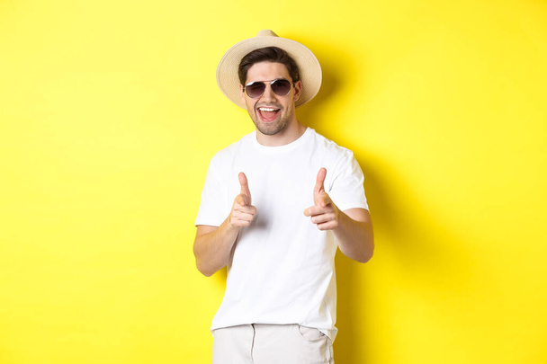 Garçon confiant et insolent en vacances flirtant avec vous, pointant du doigt la caméra et cligner des yeux, portant un chapeau d'été avec des lunettes de soleil, fond jaune - Photo, image
