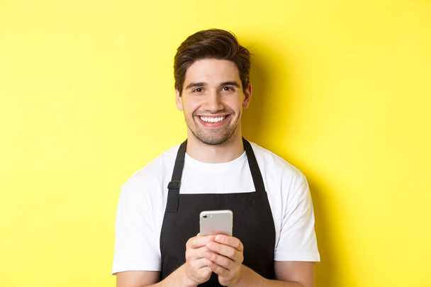 Close-up of handsome barista sending message on mobile phone, smiling happy, standing over yellow background - Foto, afbeelding