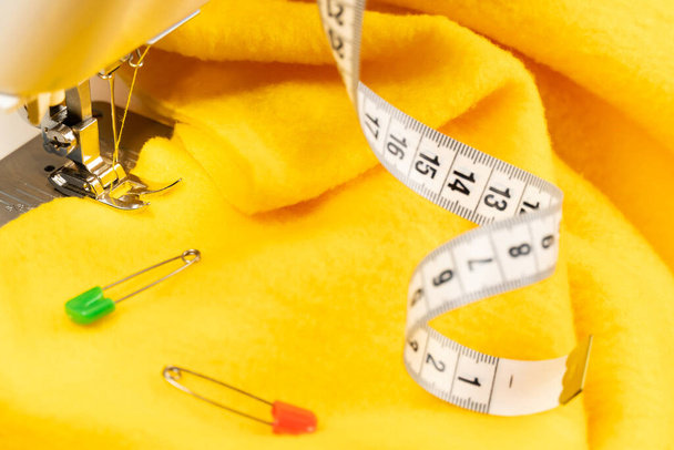 Un trozo de tela amarilla en una máquina de coser y un centímetro. La tela debajo del pie de la máquina de coser hay dos alfileres en él comienza el proceso de costura - Foto, Imagen