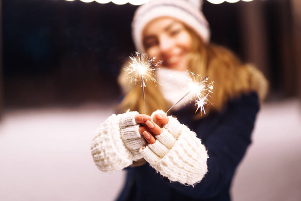 Female hand holding a burning sparkler firework bengal light.  Young woman celebrating holding sparkles in the winter forest. Festive garland lights. Christmas, New year party. - Фото, зображення