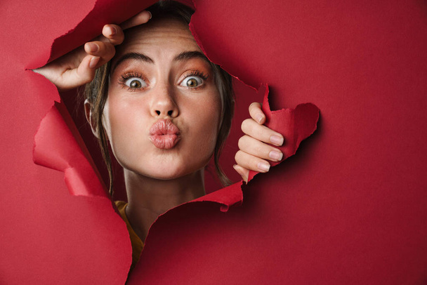 Caucásico joven feliz mujer haciendo beso cara a cámara a través de rojo fondo aislado primer plano - Foto, imagen