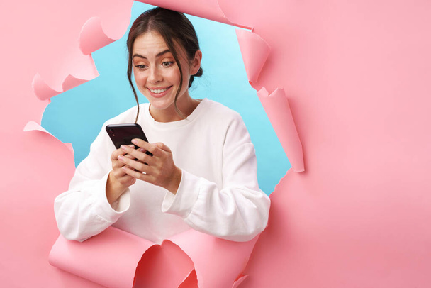 Caucasian young attractive woman smiling and holding cellphone isolated through pink background - Foto, Imagem