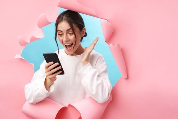 Caucasian young woman expressing surprise while holding cellphone isolated through pink background - Fotografie, Obrázek