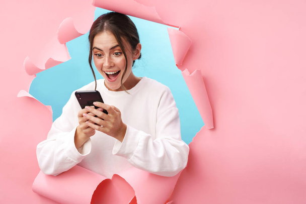 Caucasian young woman expressing surprise while holding cellphone isolated through pink background - Photo, Image