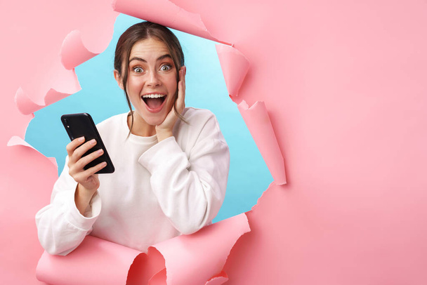 Caucasian young woman expressing surprise while holding cellphone isolated through pink background - Фото, изображение