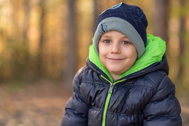 Ευτυχισμένο παιδί με καπέλο και χειμωνιάτικο μπουφάν ποζάρει μια φθινοπωρινή μέρα. Φωτογραφία στο δάσος με όμορφη bokeh. - Φωτογραφία, εικόνα
