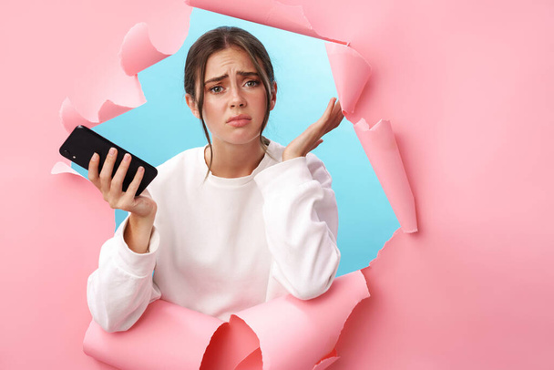 Mujer joven caucásica expresando confusión mientras sostiene el teléfono celular aislado a través de fondo rosa - Foto, imagen