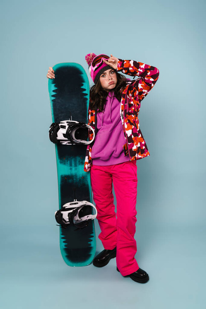 Tired young beautiful sportswoman posing with snowboard isolated over blue background - Fotografie, Obrázek