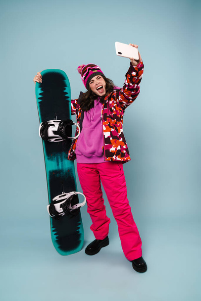 Excited sportswoman taking selfie on cellphone while posing with snowboard isolated over blue wall - Photo, image