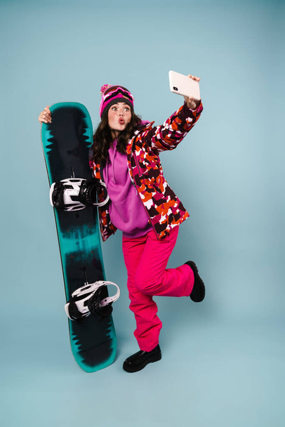 Amusing sportswoman taking selfie on cellphone while posing with snowboard isolated over blue wall - Fotografie, Obrázek