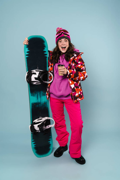 Nice excited sportswoman drinking coffee while posing with snowboard isolated over blue wall - Foto, Bild