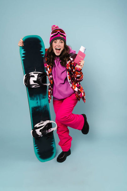 Nice excited sportswoman posing with passport and snowboard isolated over blue wall - Zdjęcie, obraz