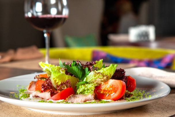 Зеленый салат с помидорами и копченой ветчиной. стакан красного вина на заднем плане - Фото, изображение