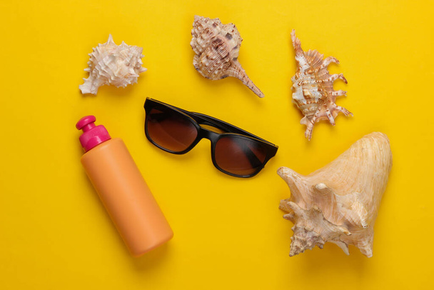 Eine Flasche Sonnencreme mit Sonnenbrille, Muscheln auf gelbem Hintergrund. Strandurlaubskonzept. Ansicht von oben - Foto, Bild