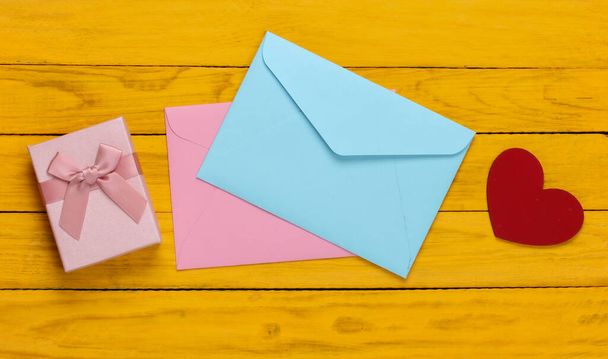ピンクと青の封筒、ギフトボックス、黄色の木製の背景にハート。バレンタインデー。トップ表示 - 写真・画像
