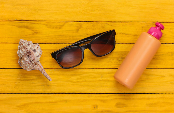 Botella de protector solar con gafas de sol, concha de mar sobre fondo de madera amarilla. Concepto de vacaciones en la playa. Vista superior - Foto, imagen