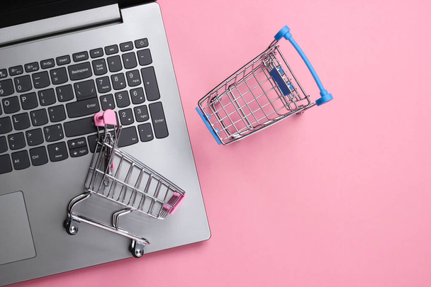オンラインショッピング。ピンクのパステルカラーの背景にスーパーマーケットトロリーノートパソコン。最上階だ。平敷 - 写真・画像