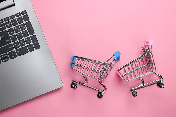 Интернет-магазины. Ноутбук с тележками супермаркета на розовом пастельном фоне. Вид сверху. Плоский лежал - Фото, изображение