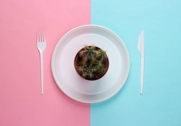 Cactus in plastic plate with disposable fork and knife on blue-pink pastel background. Top view - Photo, image