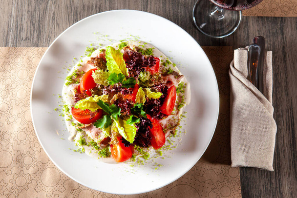 Зеленый салат с помидорами и копченой ветчиной. стакан красного вина на заднем плане. вид сверху. свет из окна ресторана - Фото, изображение