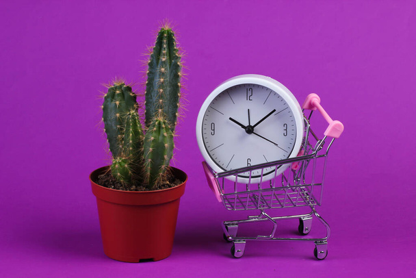 Winkeltijd. Supermarkttrolley met klok en cactus op paarse achtergrond - Foto, afbeelding