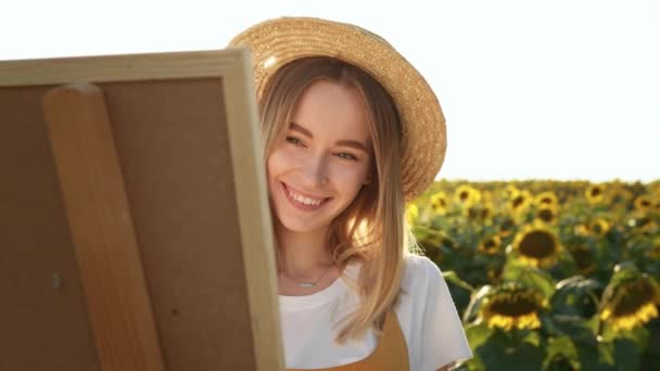 Nainen seisoo auringonkukkapellolla ja piirtää kuvan. Hän katsoo kameraan ja hymyilee. Lähikuvaa. 4K - Materiaali, video