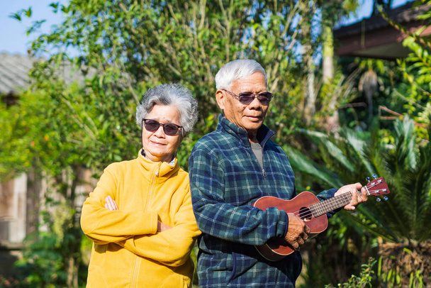 Portret romantycznego starszego Azjaty bawiącego się z żoną w ukulele, stojącego w ogrodzie. Starsza para uśmiechająca się i patrząca w kamerę. Pojęcie osób w podeszłym wieku i opieki zdrowotnej - Zdjęcie, obraz