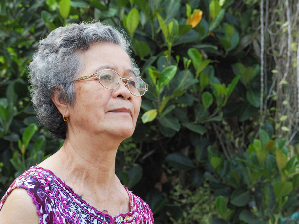 Старшая азиатка с короткими белыми волосами улыбается и смотрит в сторону, стоя в саду. Пространство для текста. Концепция пожилых людей и здравоохранения - Фото, изображение