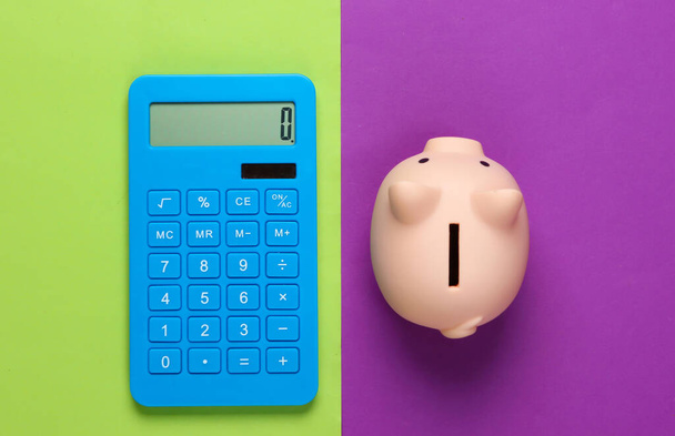Piggy bank and calculator on green purple background. Minimalistic studio shot. Overhead view. Flat lay. - Photo, Image
