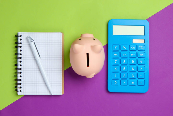 Piggy bank, notebook en rekenmachine op groene paarse achtergrond. Minimalistische studio opname. Bovenaanzicht. Vlakke plaat. - Foto, afbeelding