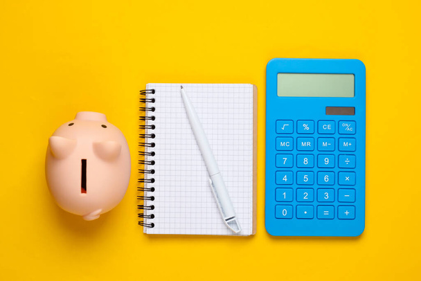 Piggy bank, notebook and calculator on yellow background. Minimalistic studio shot. Overhead view. Flat lay. - Photo, image