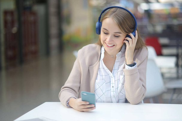 Мягкий фокус Портрет счастливой девушки, слушающей музыку онлайн с беспроводными наушниками со смартфона за белыми столами в торговом центре кафе. отдых в торговом центре. Работа вне дома - Фото, изображение