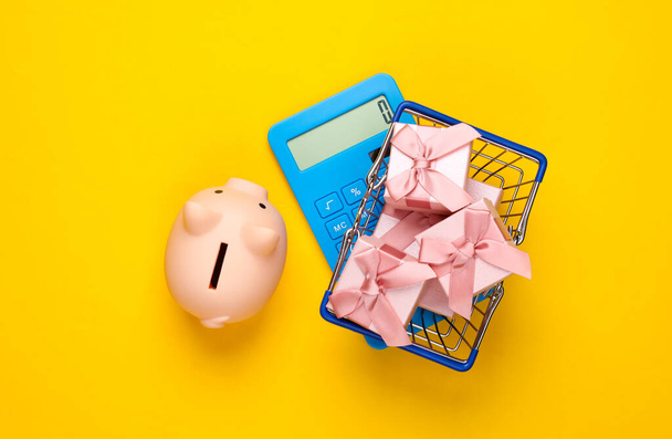 Supermarket basket with gift boxes, piggy bank and calculator on yellow background. Shopping concept. Top view - Photo, Image