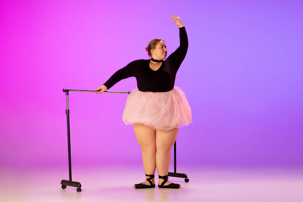 Beautiful caucasian plus size model practicing ballet dance on gradient purple-pink studio background in neon light - Foto, Imagen