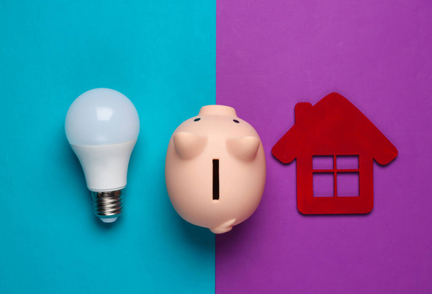 Piggy bank and led light bulb, house figure on blue purple background. Energy saving. Minimalistic studio shot. Overhead view. Flat lay. - Photo, image