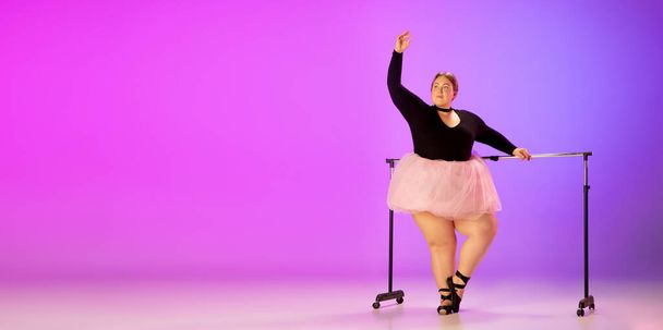 Beautiful caucasian plus size model practicing ballet dance on gradient purple-pink studio background in neon light - Фото, зображення