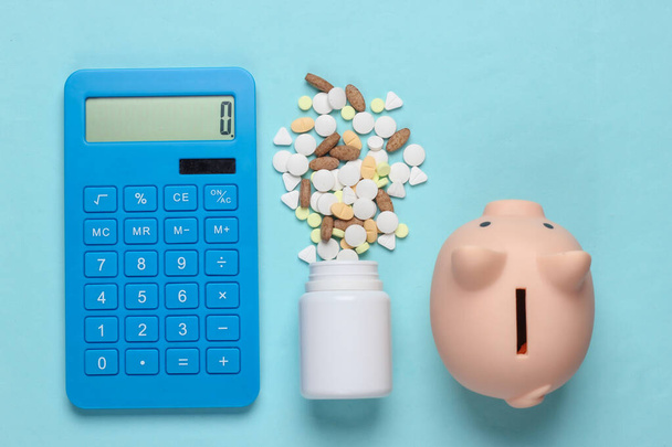 The cost of medicine. Calculator, pills, piggy bank on blue background. Top view - Фото, изображение