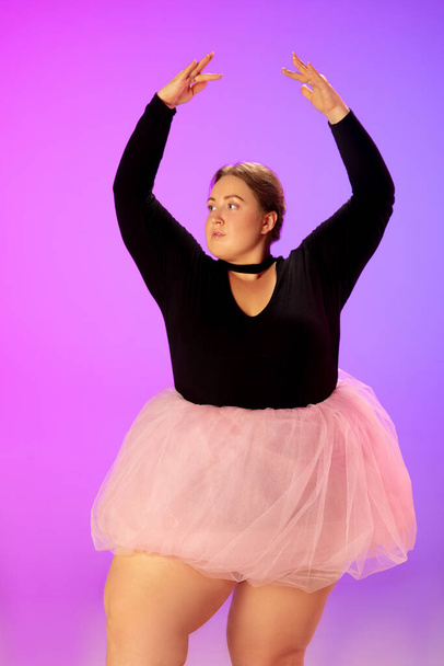 Beautiful caucasian plus size model practicing ballet dance on gradient purple-pink studio background in neon light - Foto, imagen